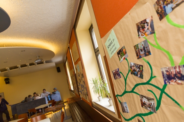 Collage an der Wand im Aufenthaltsraum, im Hintergrund sitzt eine Gruppe junger Menschen.